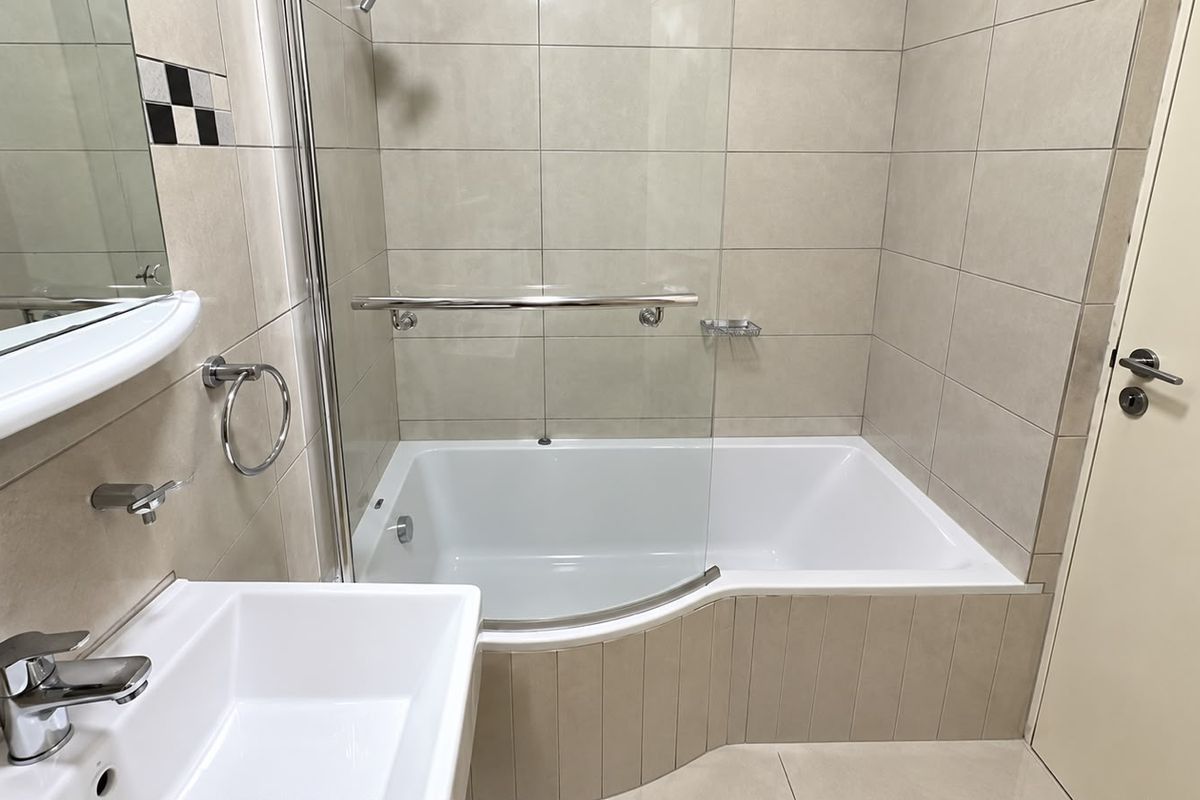 A clean, modern bathroom featuring a tiled bathtub with a glass shower screen, a wall-mounted basin, and neutral ceramic wall and floor tiles.