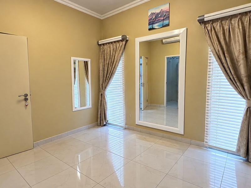 A spacious, neutral-toned bedroom featuring polished tiled flooring, large windows with venetian blinds, and integrated air conditioning.