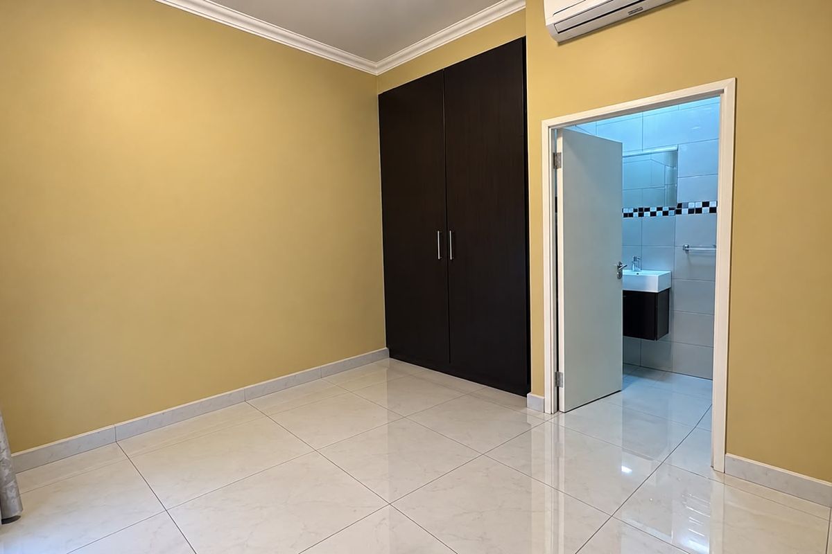 A clean, empty bedroom featuring tiled flooring, a dark wood built-in wardrobe, air conditioning, and direct access to an en-suite bathroom.
