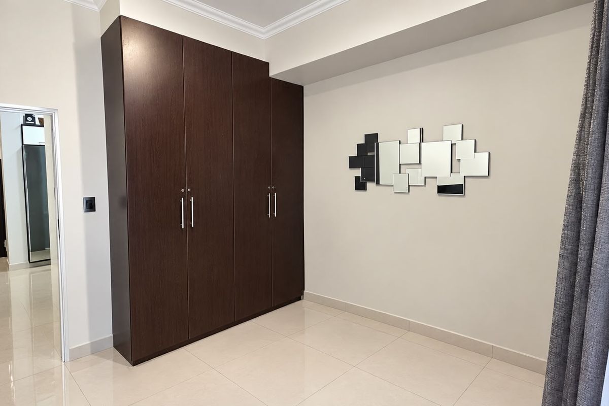 A clean, modern bedroom featuring dark wood built-in cupboards, neutral tiled flooring, and crisp white walls.