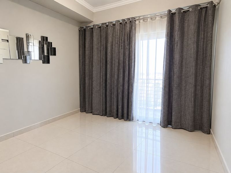 A bright, empty living room featuring polished tiled flooring, a cove ceiling, and large sliding doors leading to a balcony with floor-to-ceiling curtains.