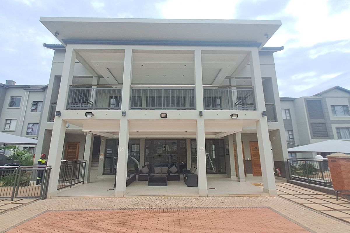 A modern apartment building exterior featuring a spacious covered ground-floor patio and an upper-level balcony with metal railings and paved outdoor areas.