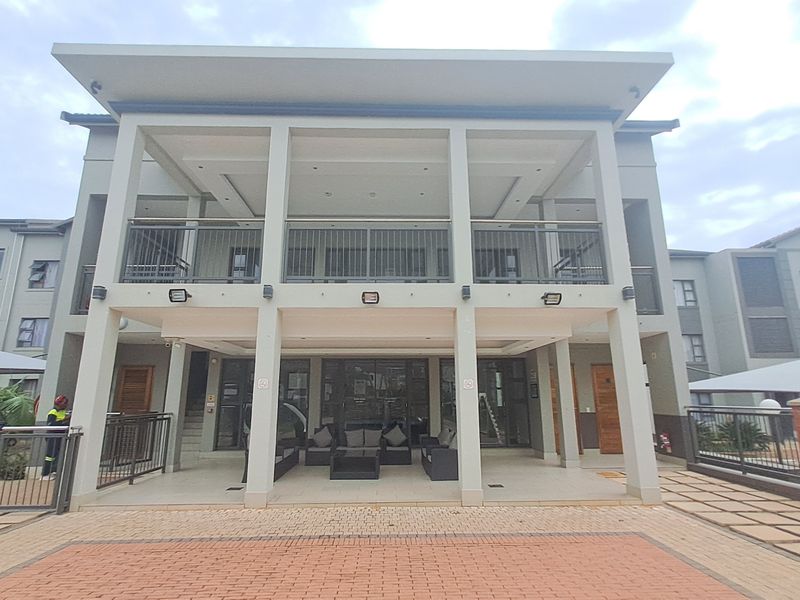 A modern apartment building exterior featuring a spacious covered ground-floor patio and an upper-level balcony with metal railings and paved outdoor areas.