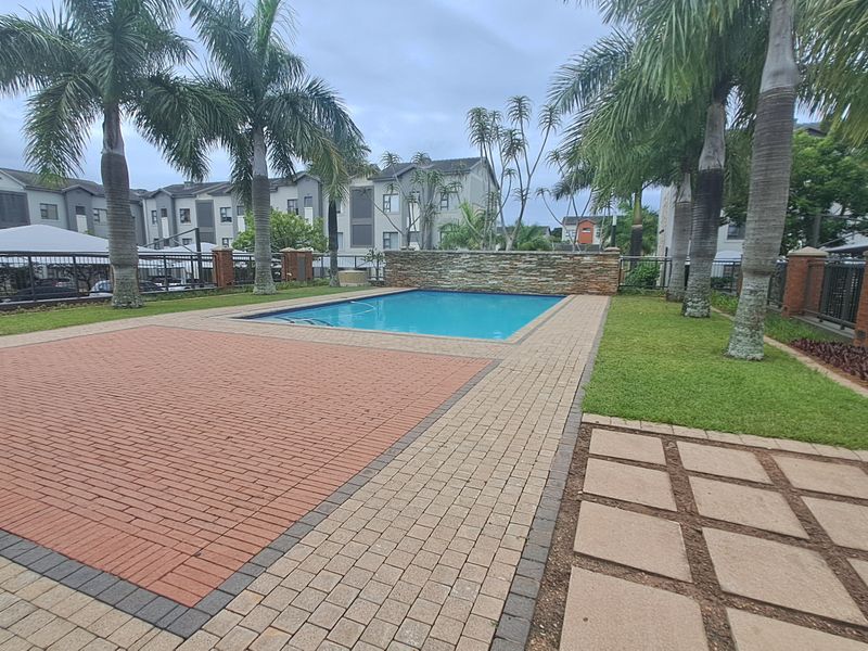 Communal pool area featuring a stone feature wall, brick paved surround, and landscaped garden within a secure apartment complex.