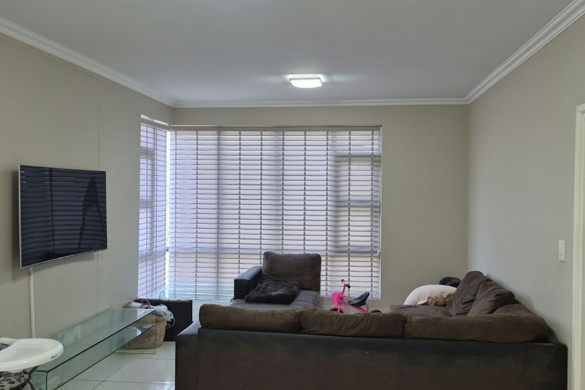 A spacious living room featuring neutral wall tones, tiled flooring, and large windows fitted with venetian blinds, providing ample natural light.