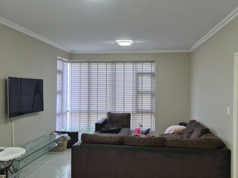 A spacious living room featuring neutral wall tones, tiled flooring, and large windows fitted with venetian blinds, providing ample natural light.