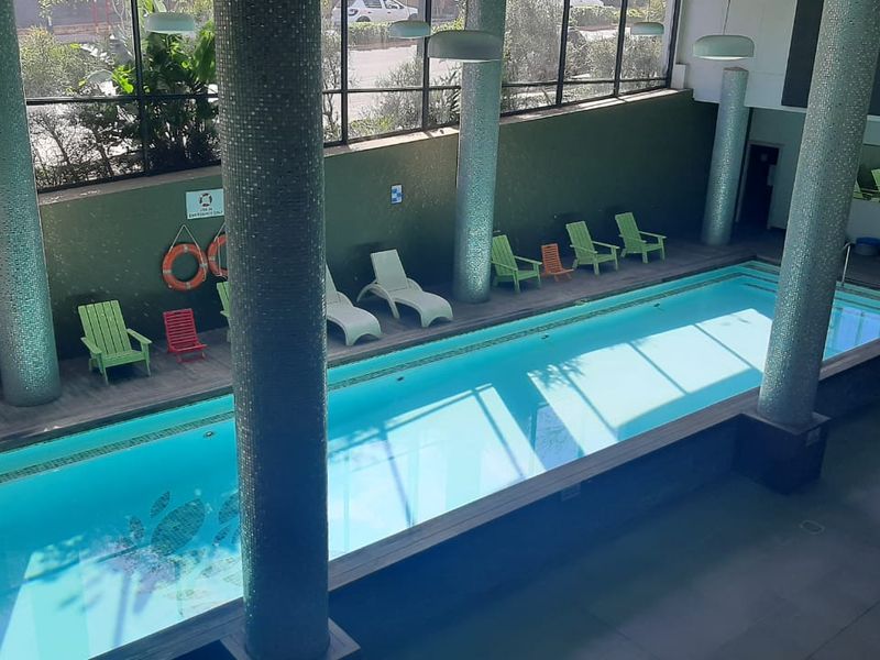 An indoor swimming pool area featuring large glass windows, mosaic-tiled structural columns, and wooden decking, providing a bright and well-maintained recreational space.
