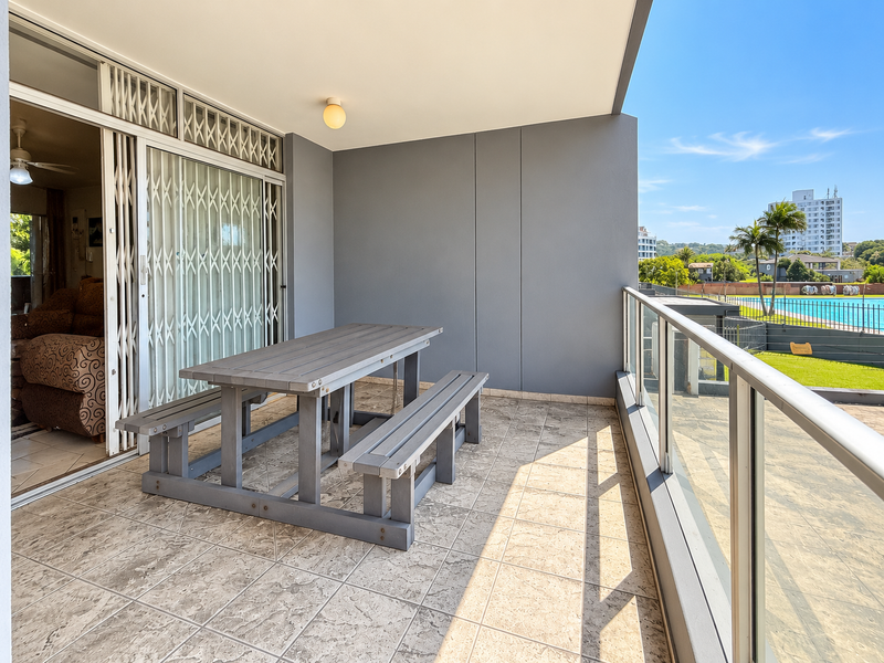 A spacious covered balcony featuring tiled flooring, a glass balustrade, and sliding door access, offering views of the surrounding complex and pool area.