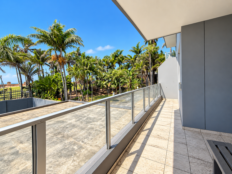 A spacious balcony featuring a modern glass balustrade and tiled flooring, offering an open outlook over the surrounding tropical garden area.