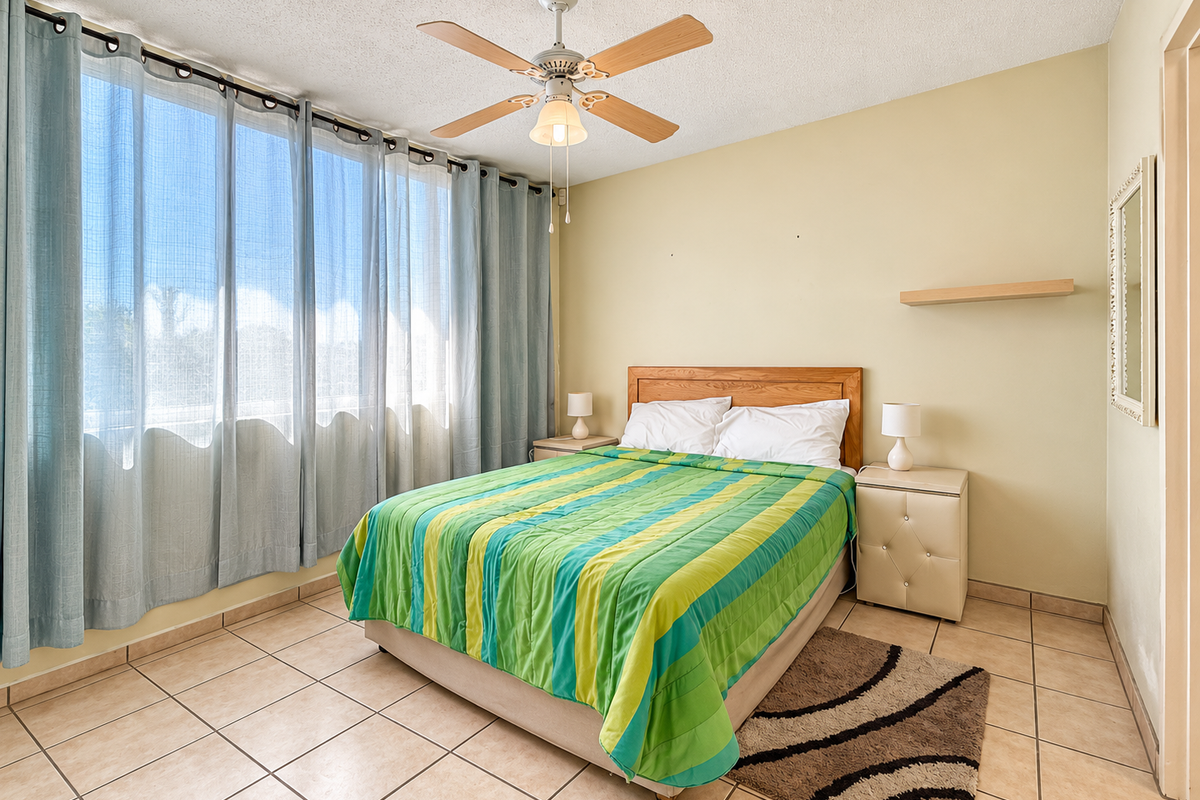A bedroom featuring tiled flooring, a ceiling fan, and a large window with natural light, providing a functional space with neutral wall tones.
