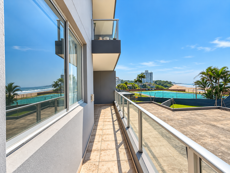 A spacious balcony featuring tiled flooring and a modern glass balustrade, offering expansive views of the pool area and the nearby coastline.