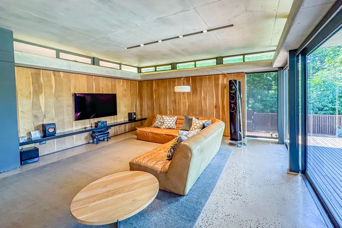 Modern living room featuring polished concrete floors, exposed concrete ceiling, warm timber wall paneling, and large sliding glass doors opening to an outdoor deck.