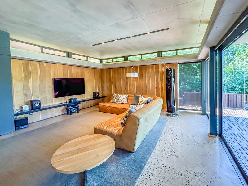 Modern living room featuring polished concrete floors, exposed concrete ceiling, warm timber wall paneling, and large sliding glass doors opening to an outdoor deck.