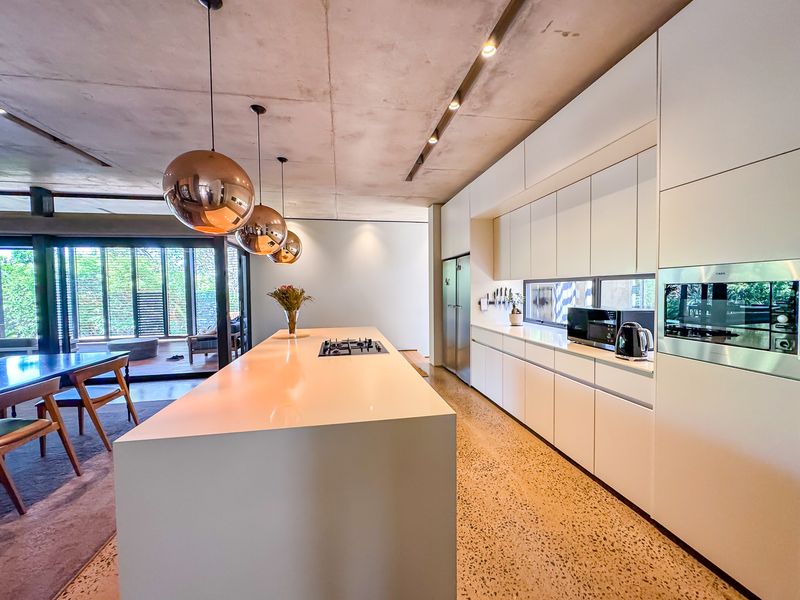 Modern open-plan kitchen featuring a large island, sleek white cabinetry, integrated appliances, polished concrete floors, and an exposed concrete ceiling with stylish pendant lighting.