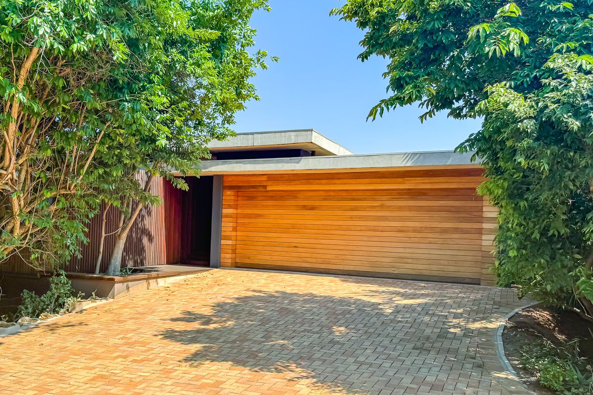 Modern front facade featuring a natural timber garage door, sleek concrete roofline, and a neatly paved driveway surrounded by mature trees.