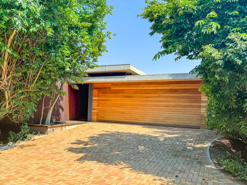 Modern front facade featuring a natural timber garage door, sleek concrete roofline, and a neatly paved driveway surrounded by mature trees.