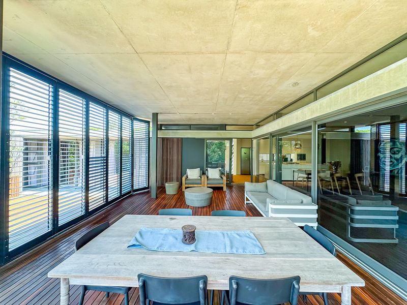 Spacious covered patio featuring timber decking, exposed concrete ceiling, and large sliding glass doors with adjustable metal louvre shutters for flexible outdoor living.