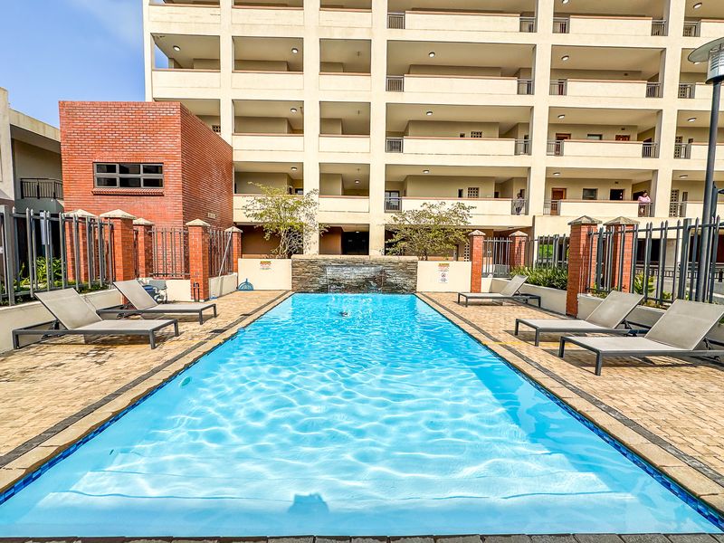 A well-maintained communal swimming pool area featuring a decorative water feature, paved decking, and secure perimeter fencing within an apartment complex.