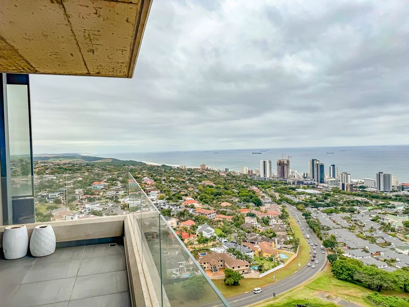 A modern balcony featuring a glass balustrade and tiled flooring, offering expansive elevated views of the surrounding coastal landscape and city skyline.