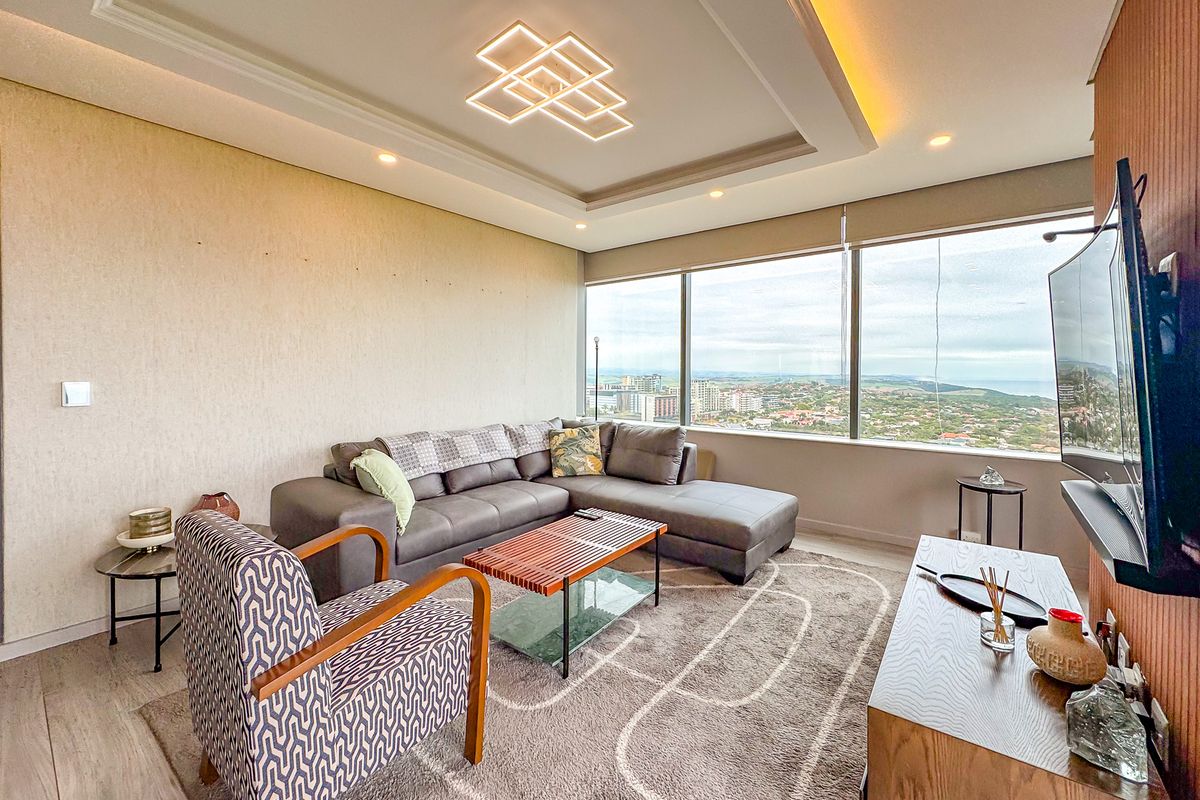 A modern living room featuring a recessed ceiling with integrated lighting, large floor-to-ceiling windows offering expansive city views, and high-quality laminate flooring.