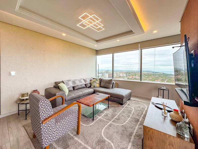 A modern living room featuring a recessed ceiling with integrated lighting, large floor-to-ceiling windows offering expansive city views, and high-quality laminate flooring.