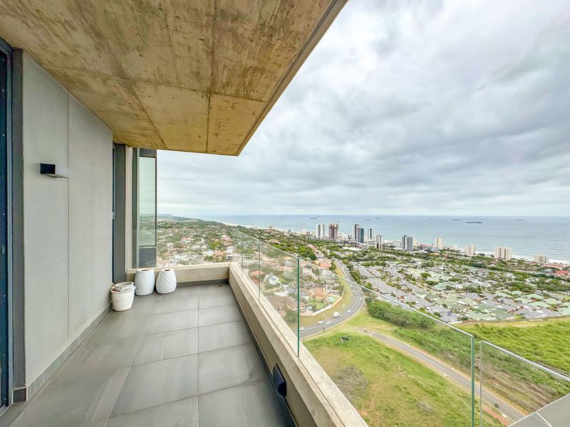 A modern, spacious balcony featuring a glass balustrade, tiled flooring, and an exposed concrete ceiling, offering expansive coastal and urban views.