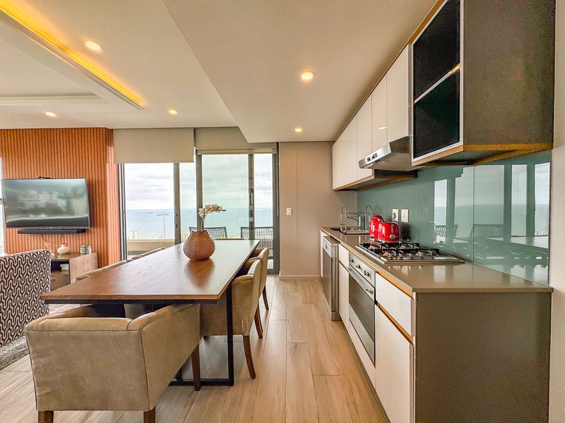 Modern open-plan kitchen and dining area featuring sleek cabinetry, stone countertops, integrated appliances, and seamless access to a balcony with ocean views.