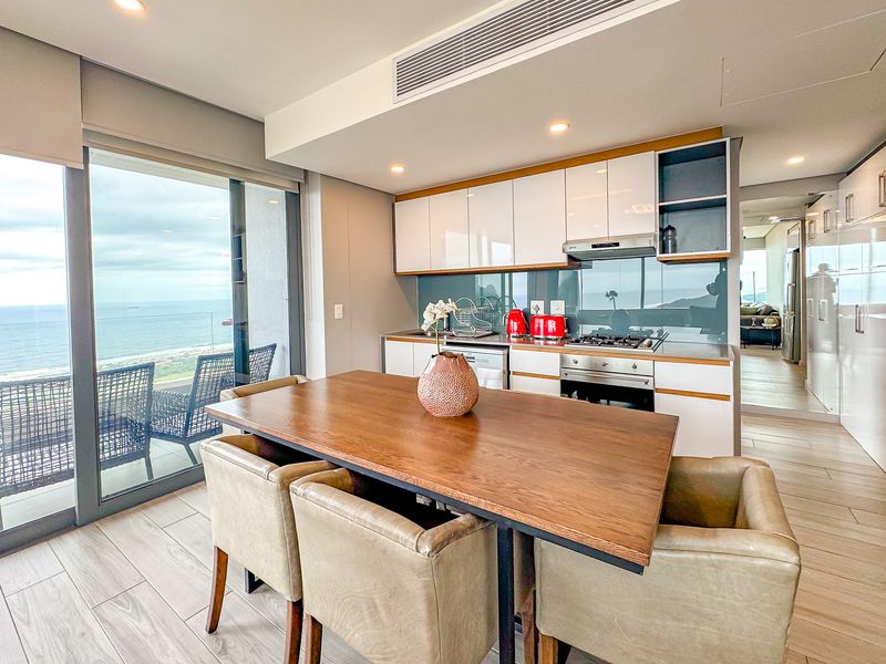 Modern open-plan kitchen and dining area featuring sleek cabinetry, integrated appliances, and large sliding doors opening to a balcony with ocean views.