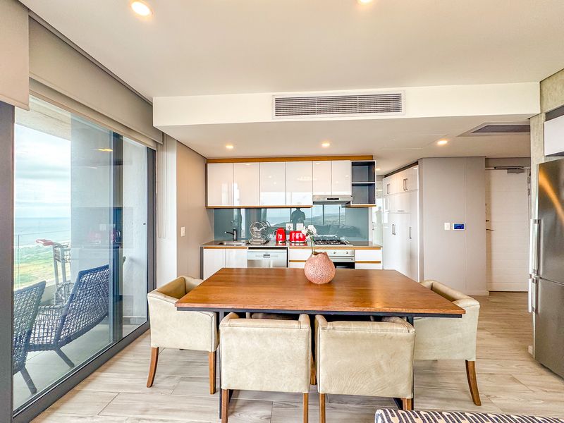 Modern open-plan kitchen and dining area featuring sleek cabinetry, integrated appliances, ducted air conditioning, and large sliding doors leading to a balcony with views.