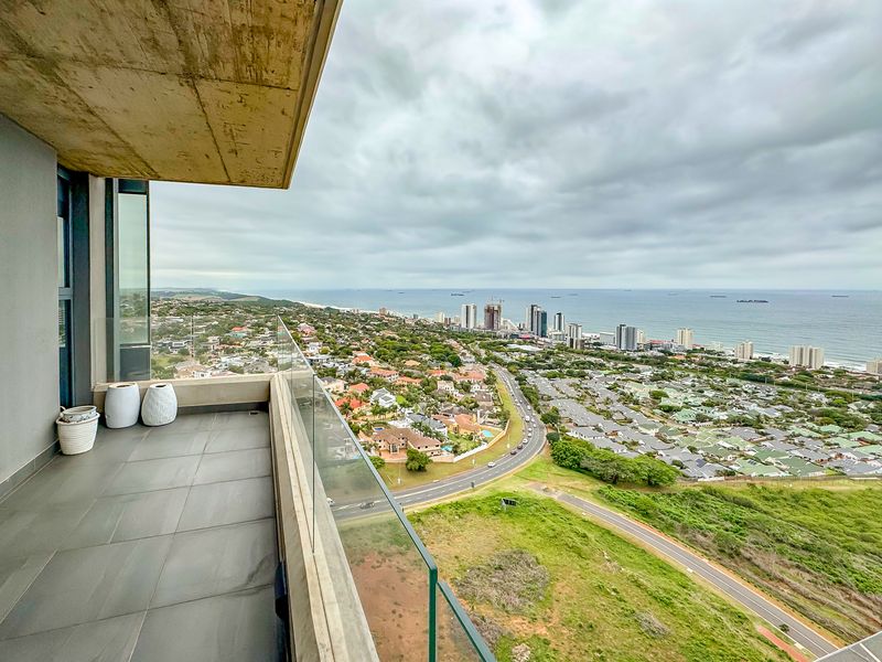 A modern balcony featuring a glass balustrade and tiled flooring, offering expansive elevated views of the surrounding coastal landscape and city skyline.