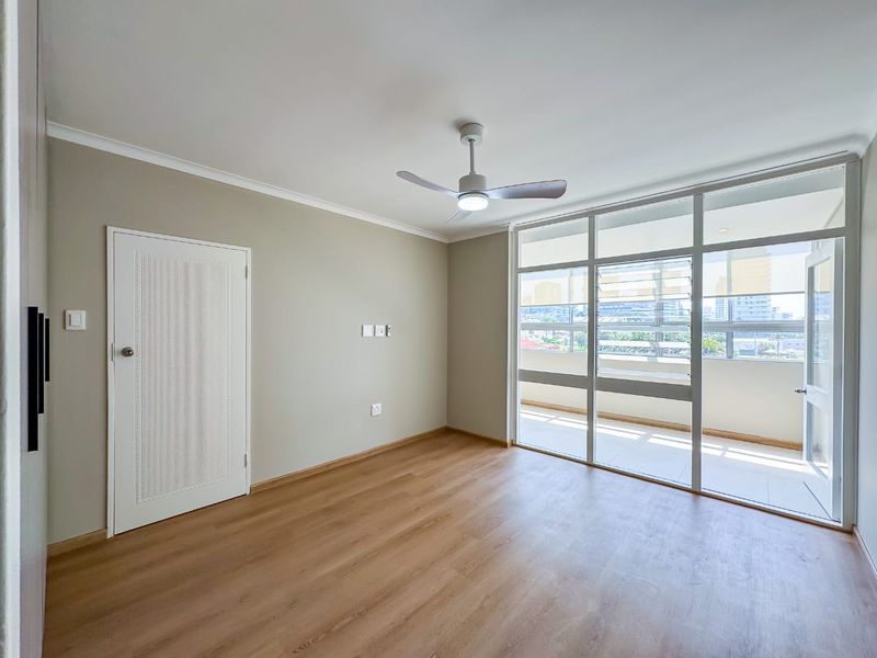 A bright, empty bedroom featuring modern laminate flooring, a ceiling fan, and a large glass door providing access to a private balcony.