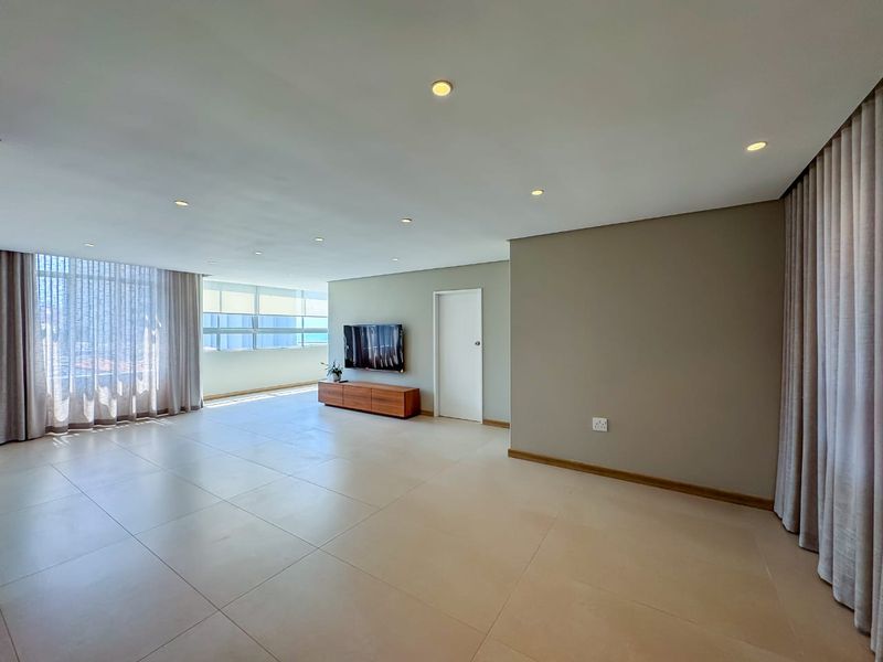 Spacious, light-filled living area featuring neutral tiled flooring, recessed downlighting, and large windows with floor-to-ceiling curtains.
