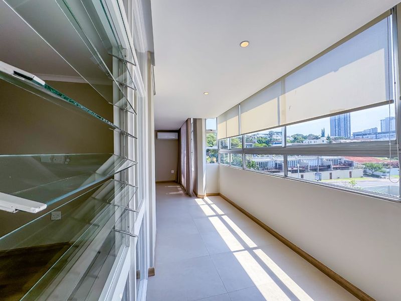 A bright, enclosed balcony featuring tiled flooring, louvered glass windows, and a wall-mounted air conditioning unit, offering an open and versatile space.