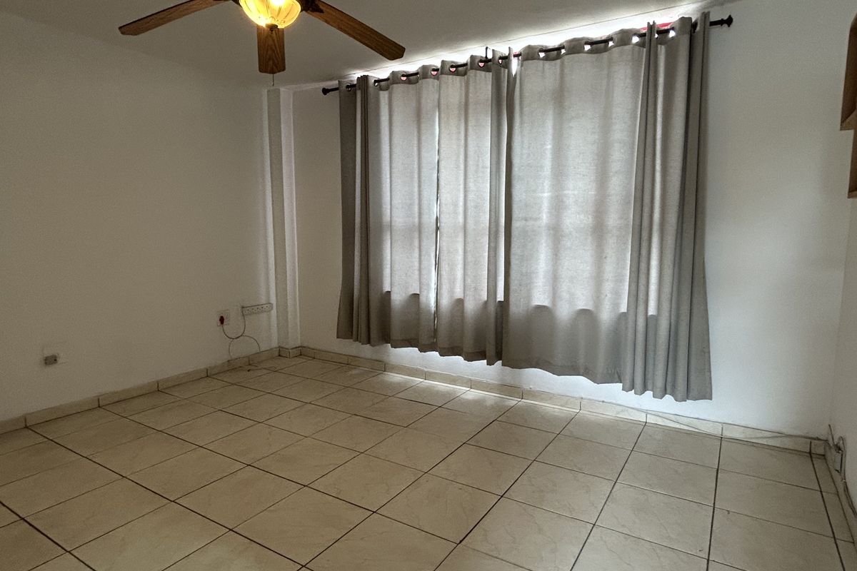 A bright, empty living room featuring neutral tiled flooring, a ceiling fan, and large windows with curtains, offering a blank canvas for personalization.
