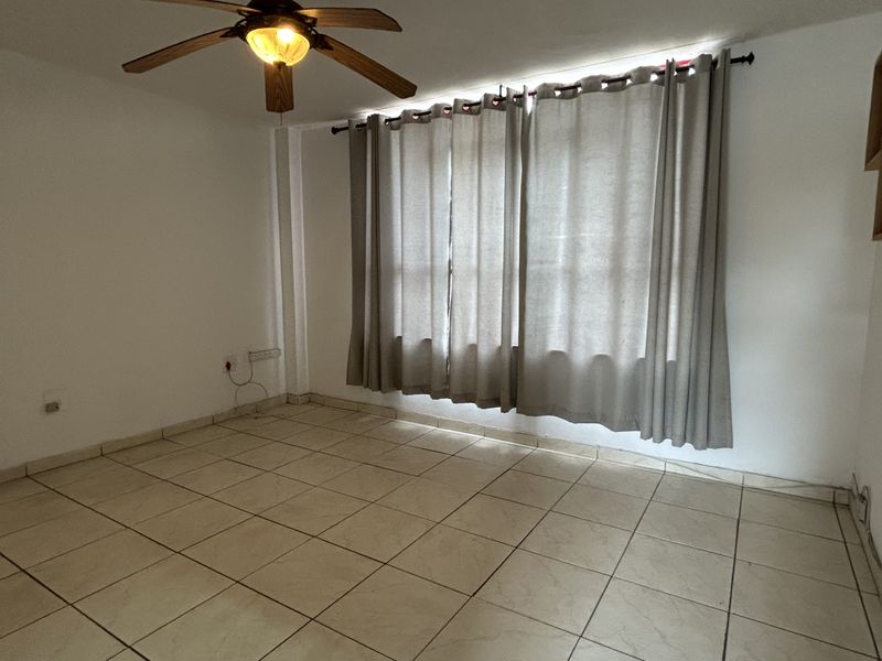 A bright, empty living room featuring neutral tiled flooring, a ceiling fan, and large windows with curtains, offering a blank canvas for personalization.