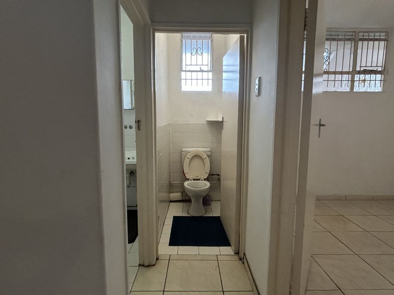 A functional bathroom featuring ceramic floor and wall tiles, a standard toilet, and a window fitted with security bars.