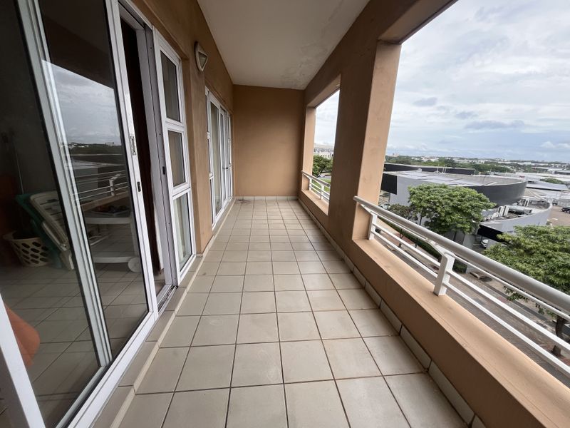 A spacious covered balcony featuring tiled flooring, metal railings, and sliding glass door access, offering an elevated view of the surrounding area.