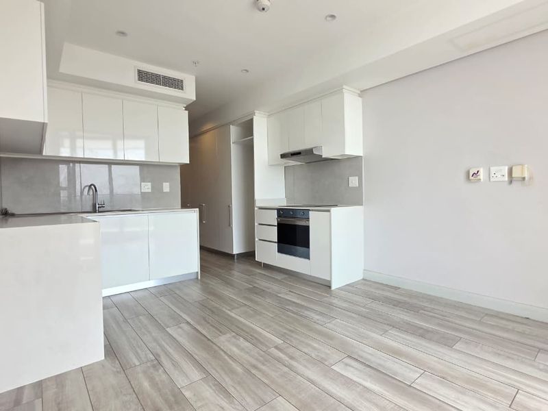 A modern, open-plan kitchen and living area featuring sleek white cabinetry, integrated appliances, and light wood-look tiled flooring in excellent condition.