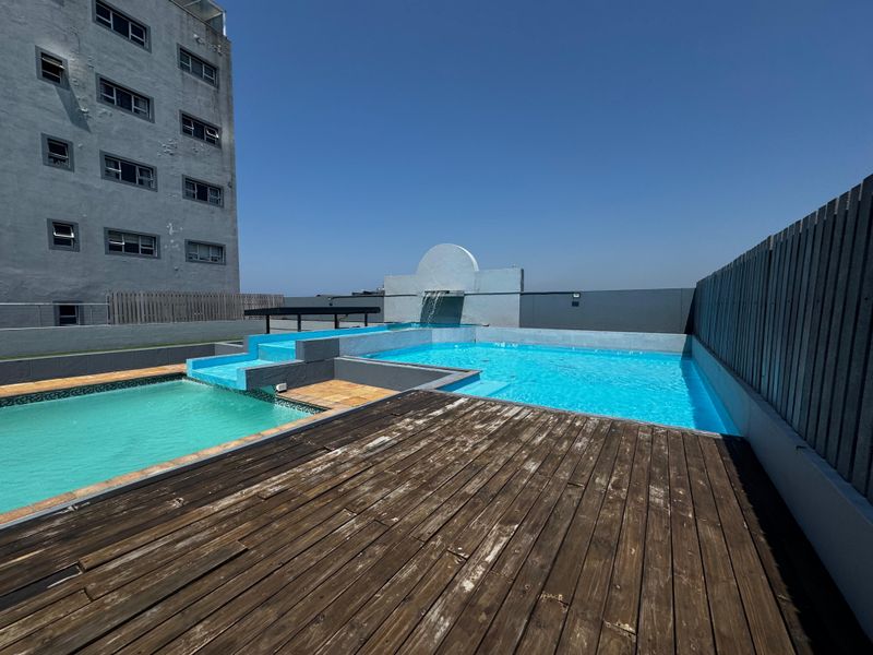 Rooftop swimming pool area featuring a water feature, tiled pool surrounds, and timber decking with views of the adjacent building facade.