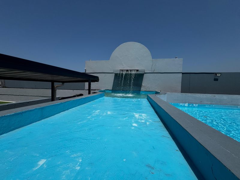 An outdoor swimming pool area featuring a built-in water feature and a metal pergola structure, set against a clear blue sky.