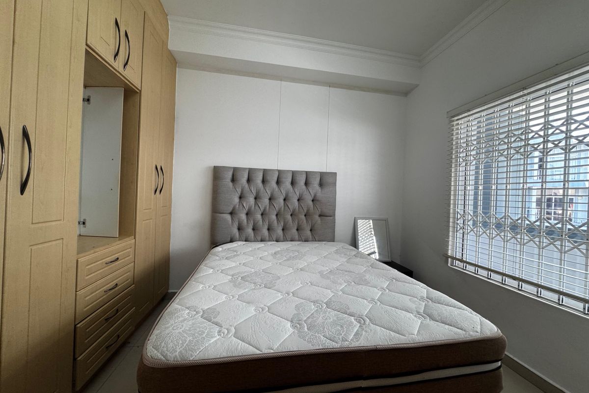 A well-maintained bedroom featuring built-in wooden cupboards, neutral finishes, and a window fitted with security gates and venetian blinds.
