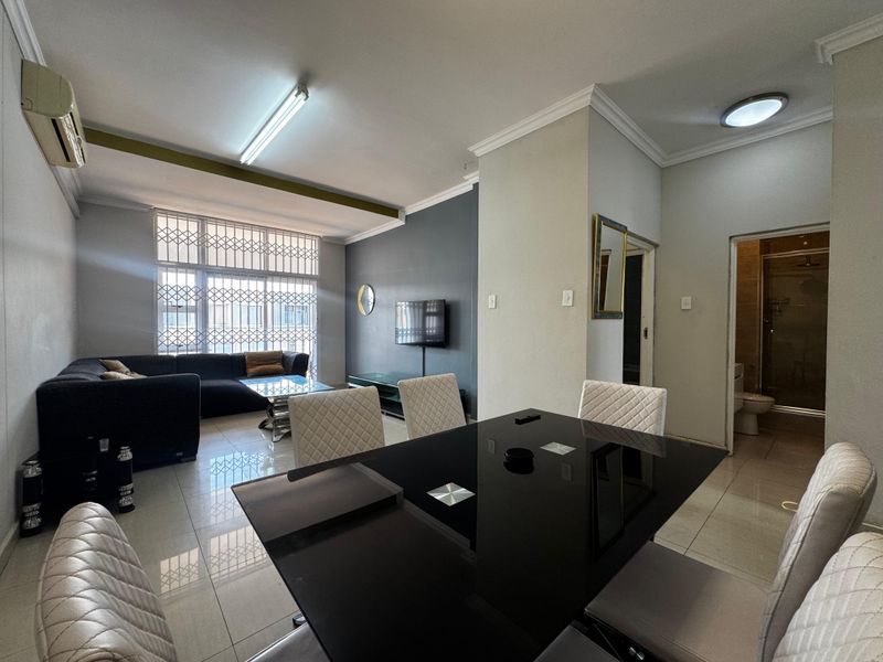 An open-plan living and dining area featuring tiled floors, modern lighting, and a wall-mounted air conditioning unit, with direct access to an adjacent bathroom.