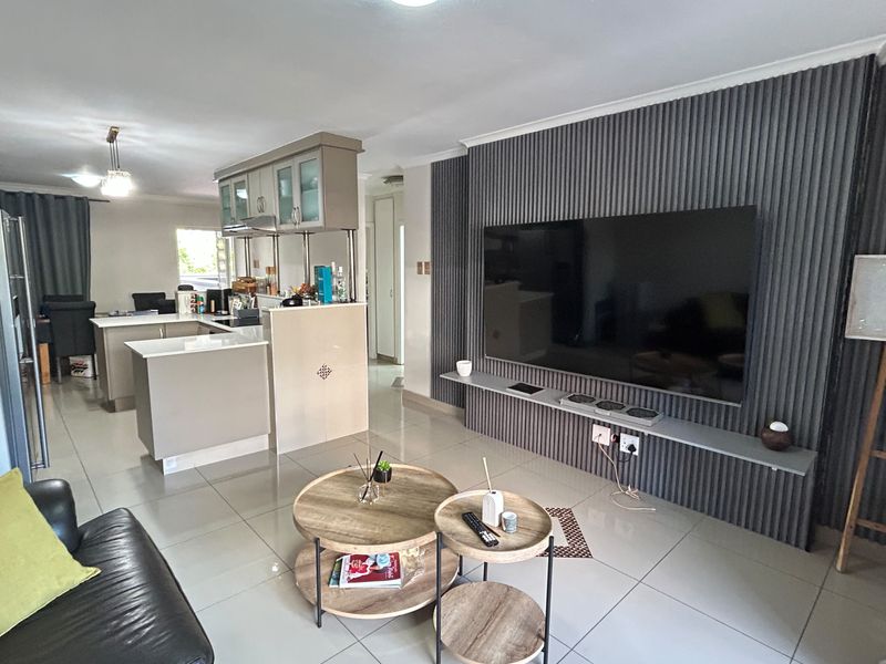 A modern open-plan living and kitchen area featuring tiled floors, a stylish grey slatted feature wall with a floating shelf, and a functional kitchen island.