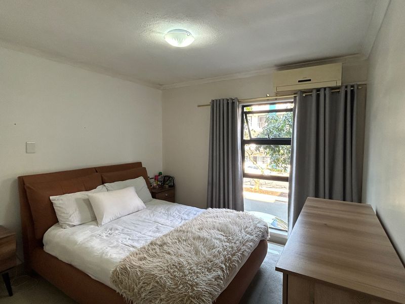 A well-lit bedroom featuring a wall-mounted air conditioning unit and a large window with curtains, providing natural light and ventilation.
