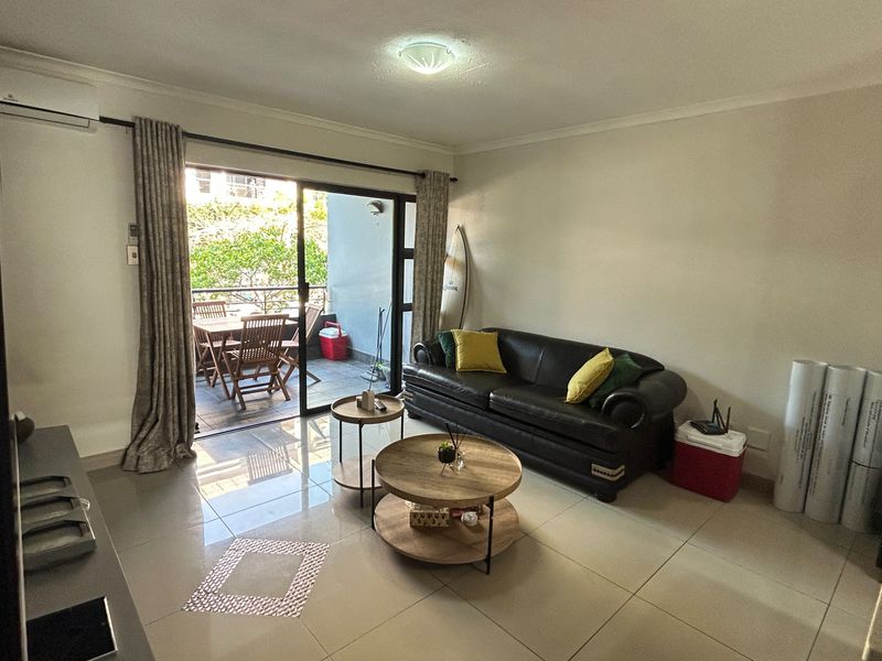 A bright living room featuring large sliding glass doors that open onto a private balcony, complemented by neutral tiled flooring and air conditioning.
