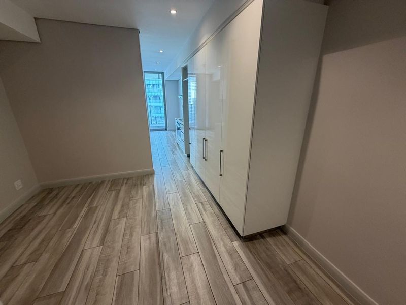 Modern open-plan living area featuring sleek white built-in cabinetry, recessed downlighting, and contemporary wood-look tile flooring leading to a balcony.
