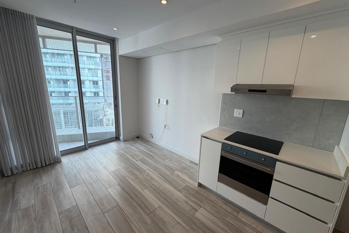 Modern open-plan kitchen and living area featuring sleek white cabinetry, integrated appliances, wood-look tile flooring, and large sliding doors leading to a balcony.