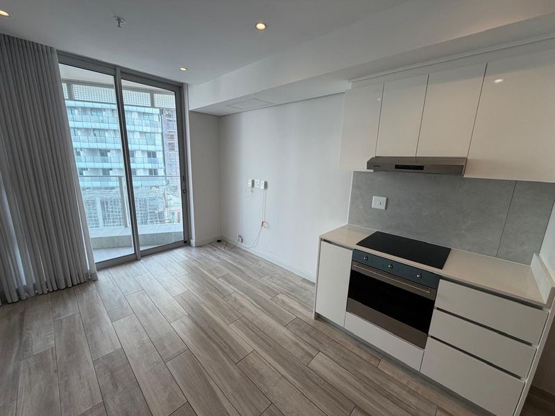 Modern open-plan kitchen and living area featuring sleek white cabinetry, integrated appliances, wood-look tile flooring, and large sliding doors leading to a balcony.
