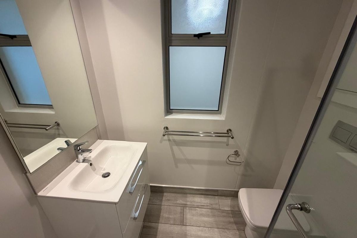 A modern, clean bathroom featuring a sleek vanity, wall-hung toilet, shower enclosure, and contemporary wood-look tiled flooring with ample natural light.