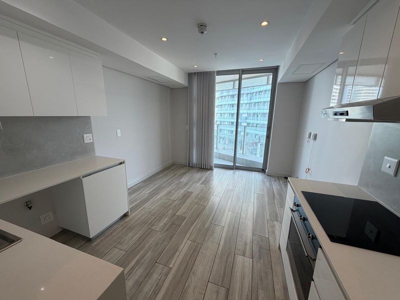 Modern open-plan kitchen and living area featuring sleek white cabinetry, stone countertops, integrated appliances, and wood-look tile flooring with access to a private balcony.